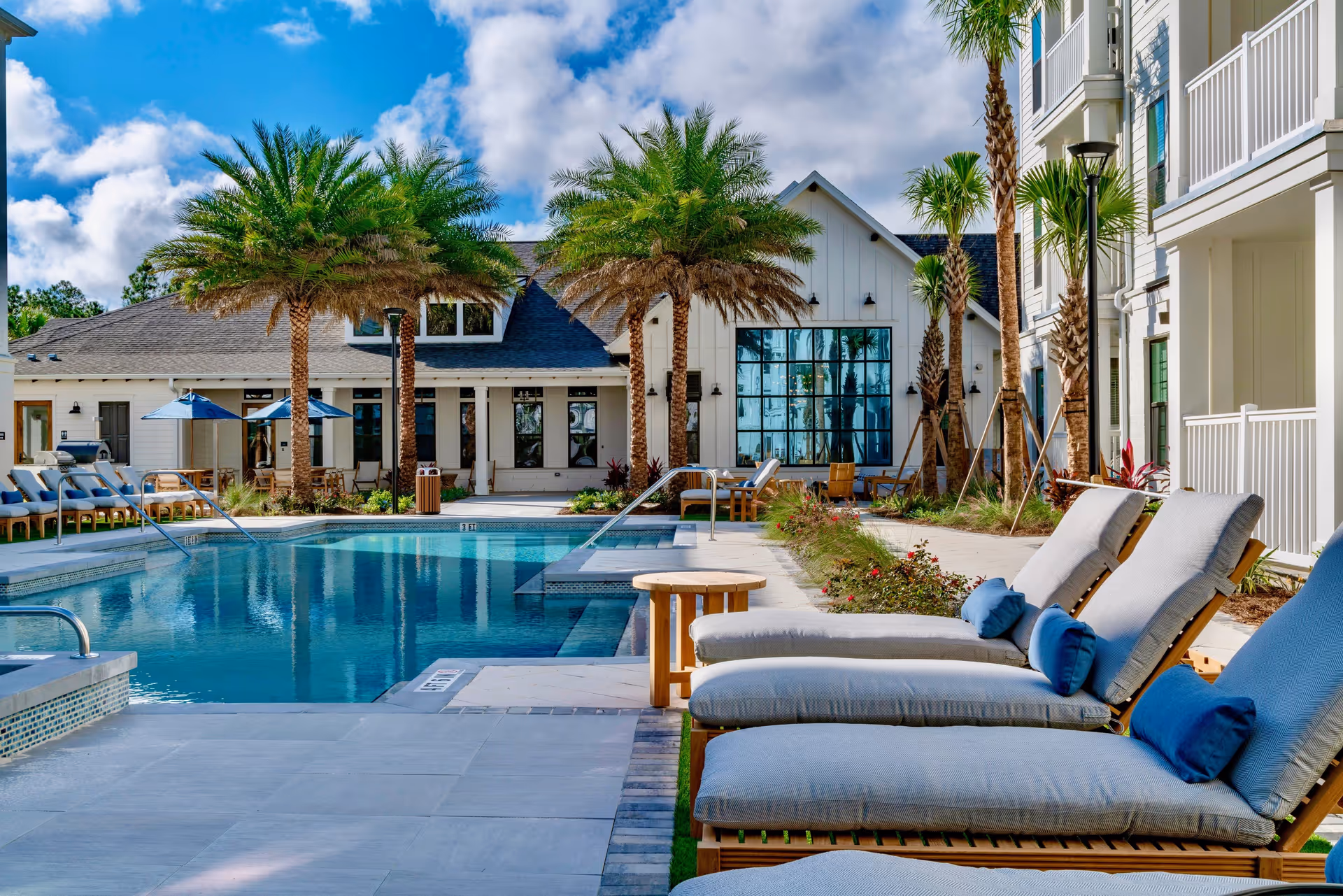 Outdoor swimming pool area at a senior living facility with lounge chairs featuring blue cushions and pillows, palm trees, umbrellas, and a white building with large windows in the background under a partly cloudy sky.