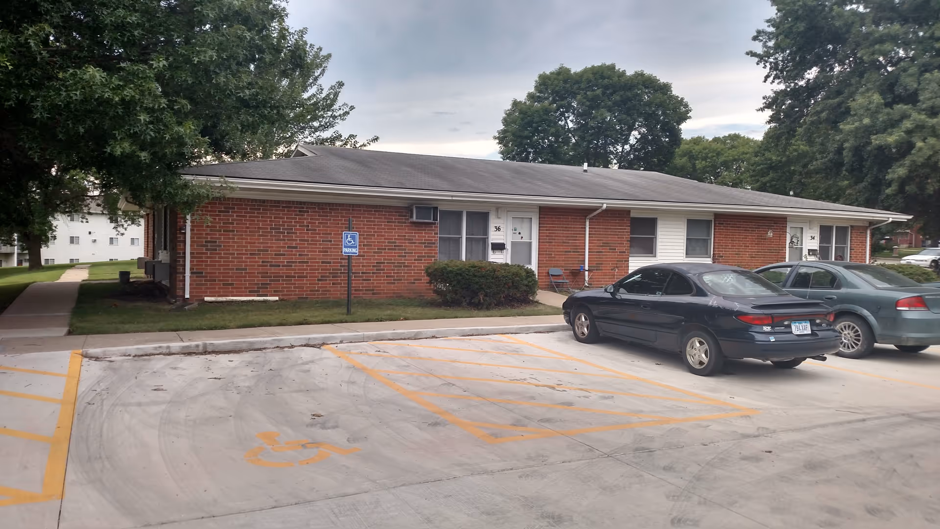 Low single-story brick apartment building with a parking lot, two parked cars, and a visible handicap parking sign.