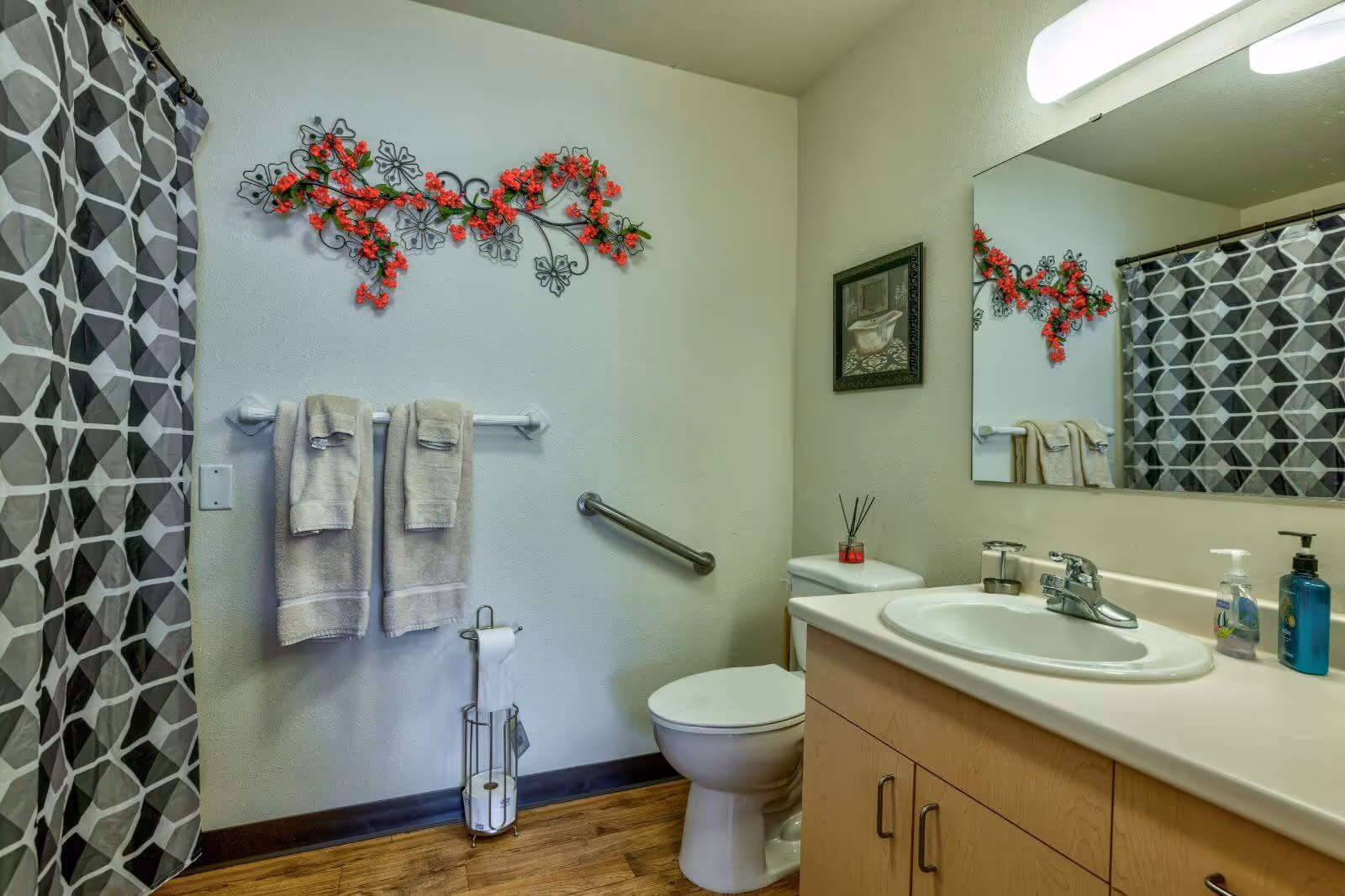 A bathroom with a white toilet, a sink with a countertop and wooden cabinets, a large mirror above the sink, a towel rack with two beige towels, a toilet paper holder with extra rolls, a shower curtain with a geometric pattern, and decorative wall art featuring red flowers and a framed picture.
