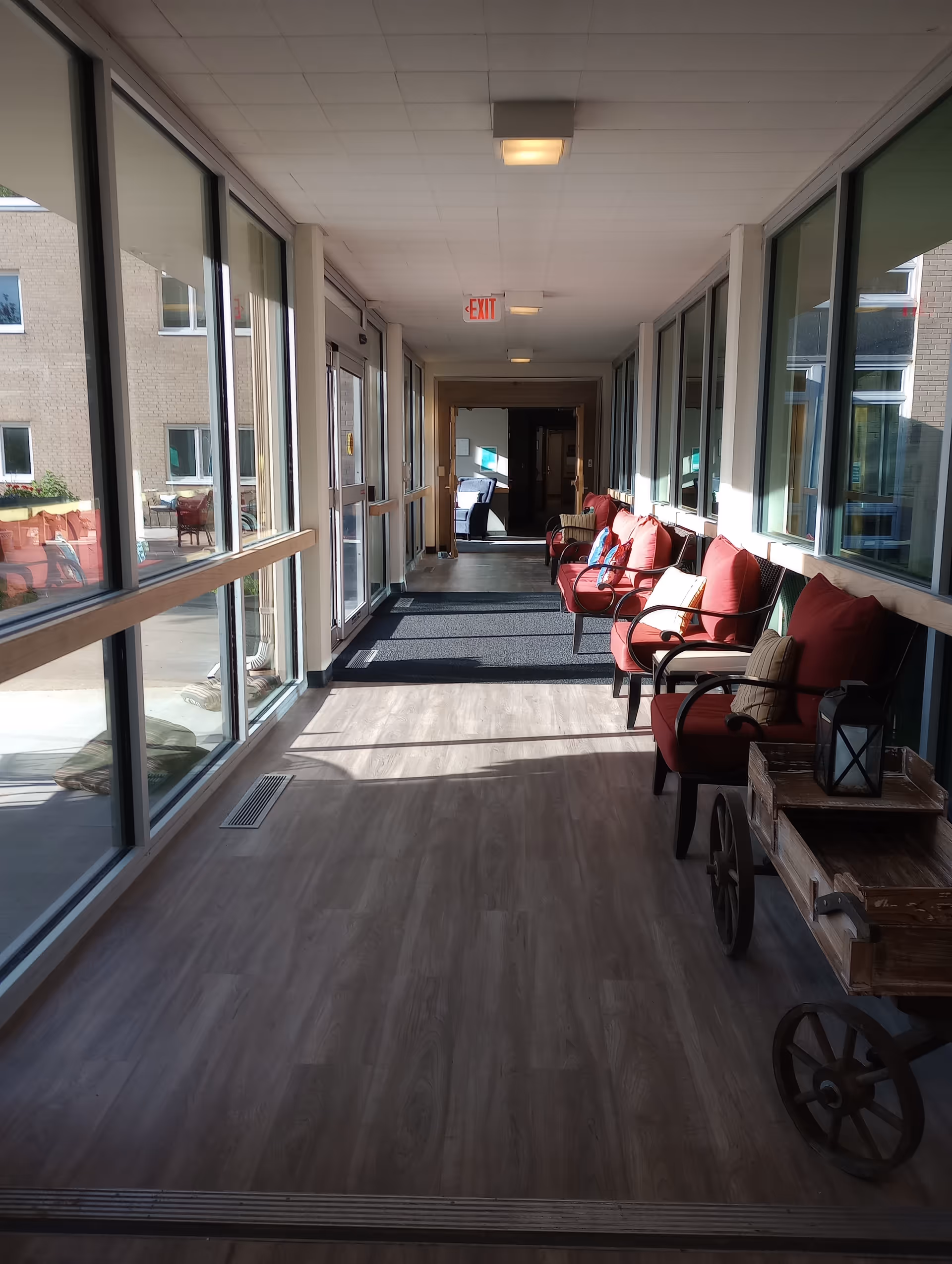 A bright indoor hallway with large windows on both sides allowing natural light to fill the space. On the right side, there are several red cushioned chairs with pillows arranged along the wall. A wooden cart with decorative items is also visible. The floor is covered with light wood-patterned flooring, and there is a dark mat near the exit door at the end of the hallway.