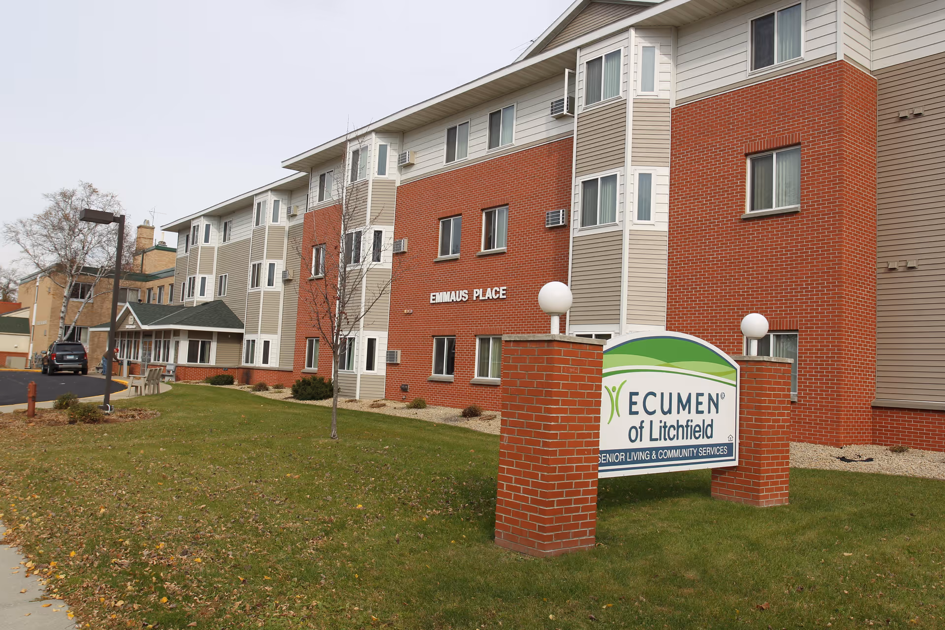 Exterior view of a senior living facility named Ecumen of Litchfield. The building has a combination of red brick and beige siding with multiple windows. A sign in front of the building reads 'Ecumen of Litchfield Senior Living & Community Services.' There is a grassy area with a sidewalk and a few small trees in front of the building.