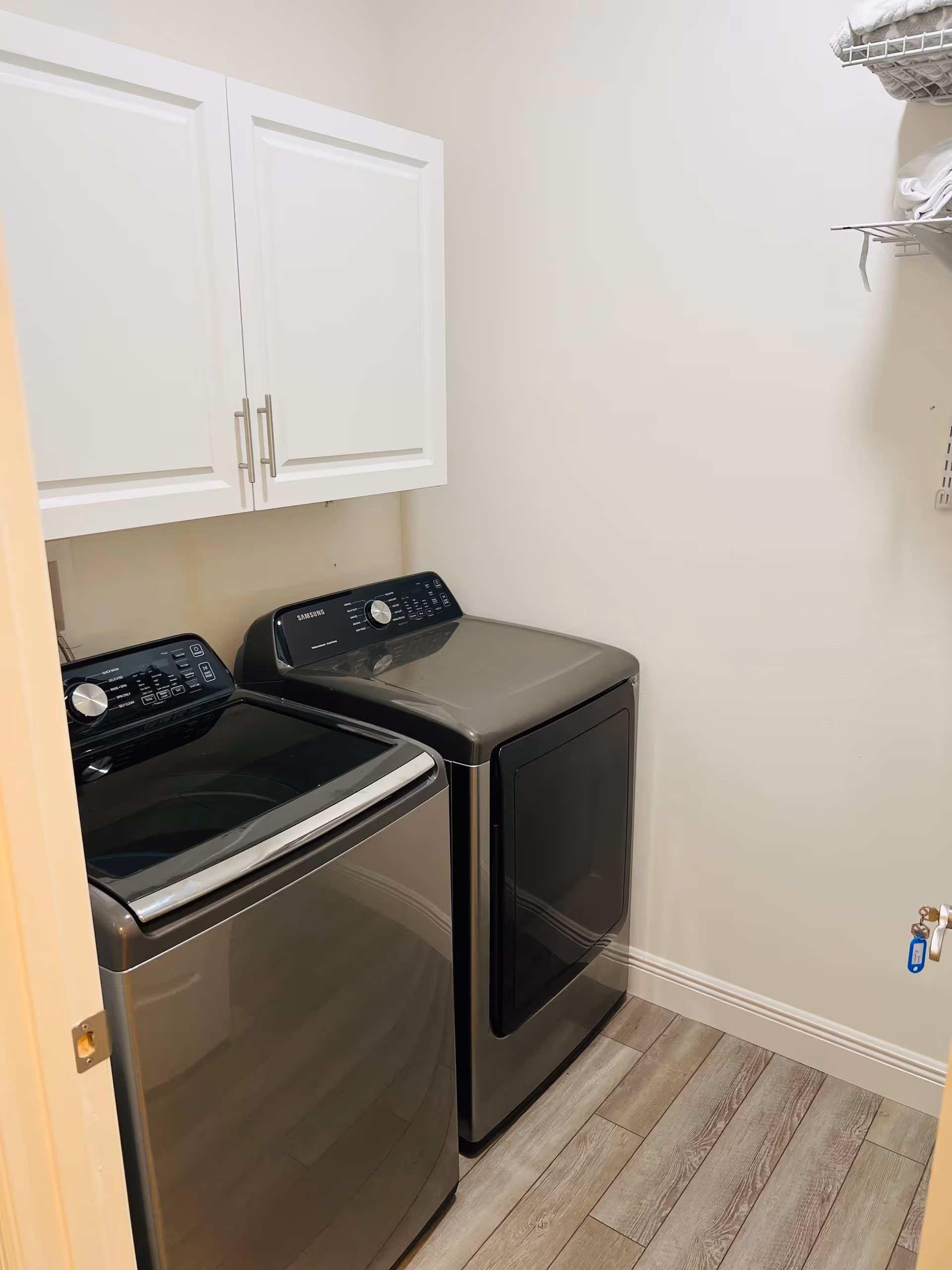 A small laundry room with a modern black Samsung washing machine and dryer side by side. Above the machines are white cabinets for storage, and to the right is a wire shelf holding folded towels. The floor has wood-like tiles and the walls are painted white.