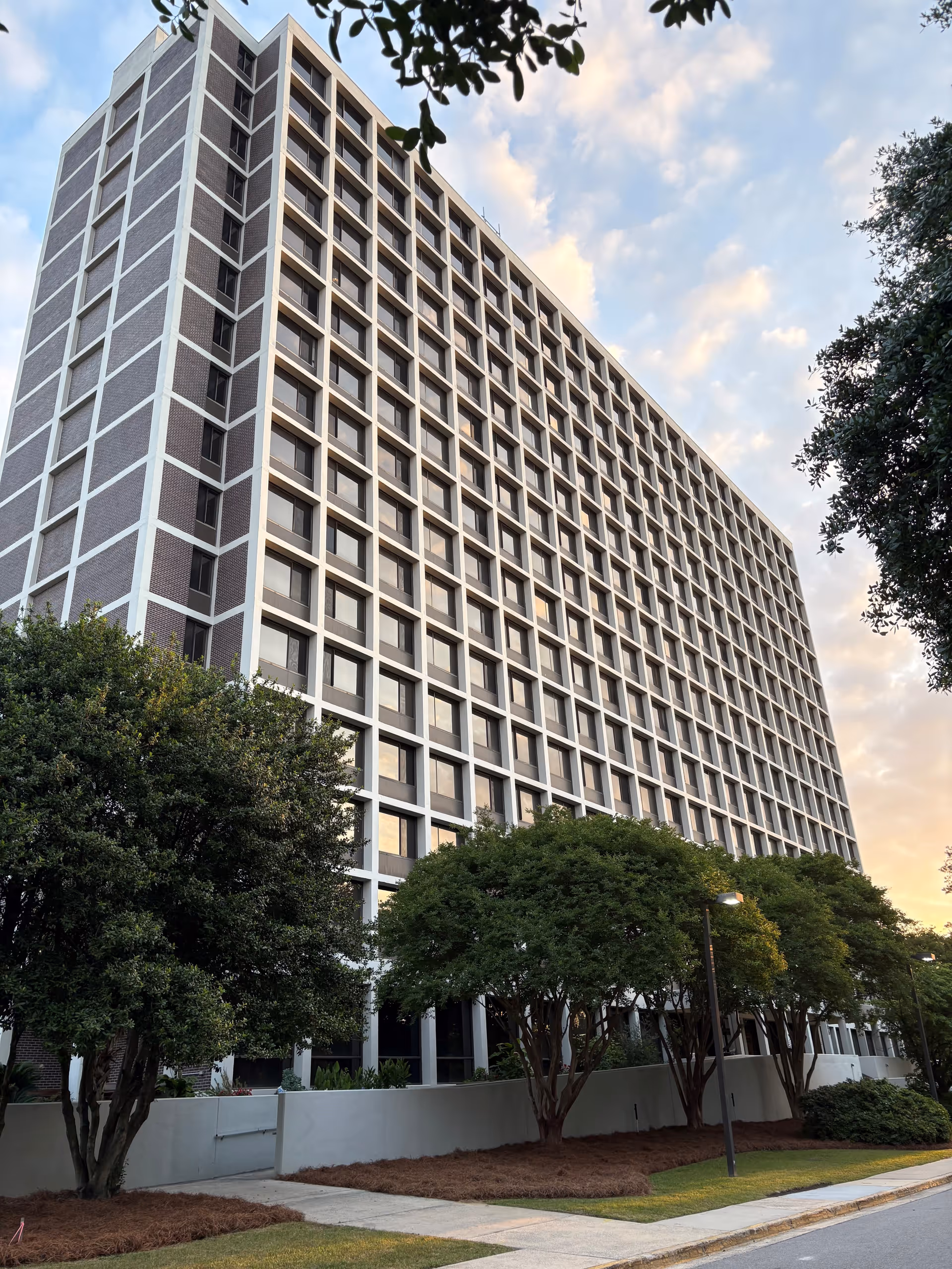 Tall multi-story building with a grid-like pattern of windows, surrounded by trees and a sidewalk, under a partly cloudy sky during sunset.