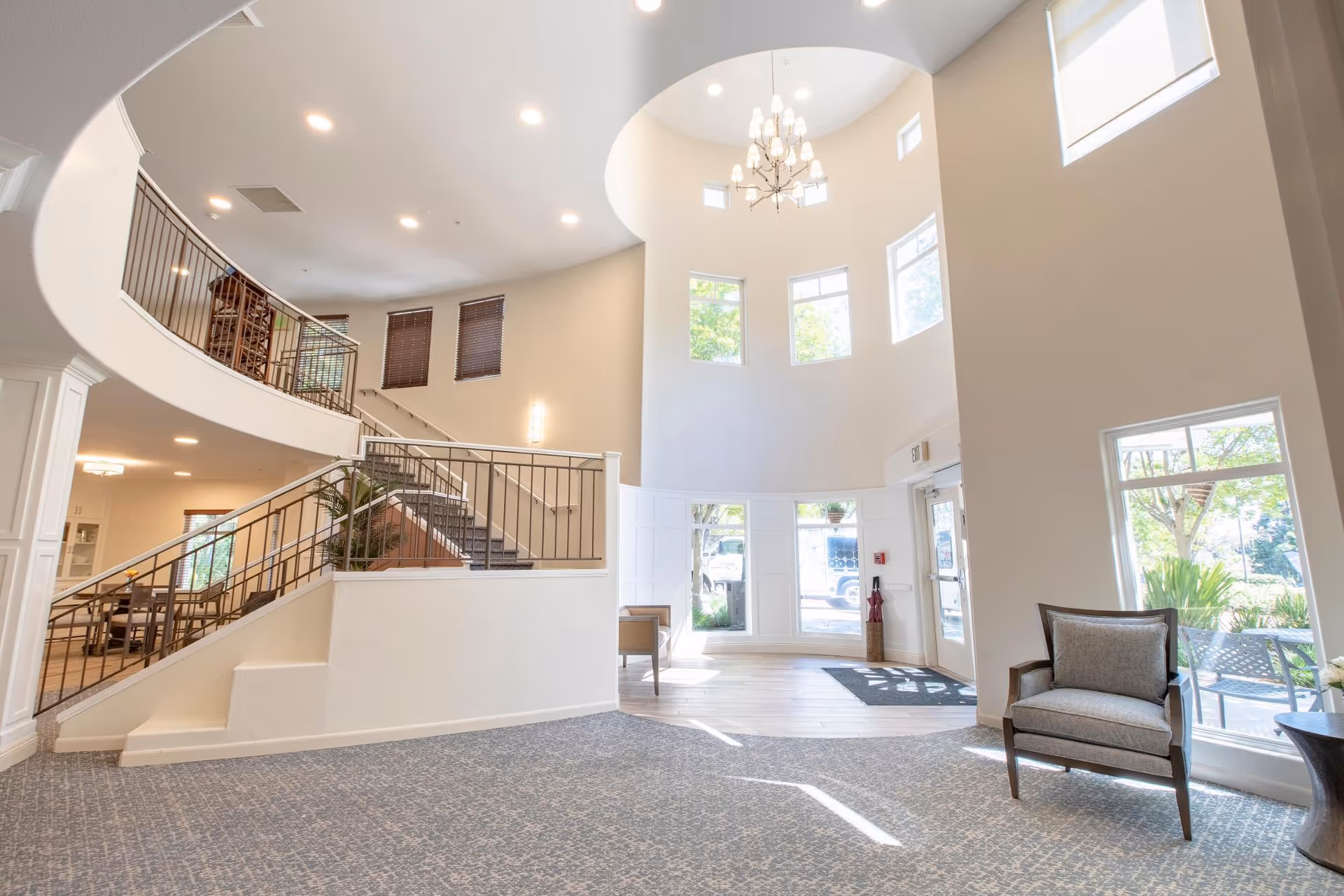 Bright and spacious interior of a senior living facility lobby with a high ceiling, large windows letting in natural light, a chandelier, a staircase with metal railings, and a single armchair near the entrance door.