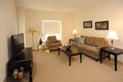 A cozy living room with a beige sofa, an armchair, a black coffee table, two side tables with lamps, a floor lamp, a flat-screen TV on a black TV stand, and two framed pictures on the wall.