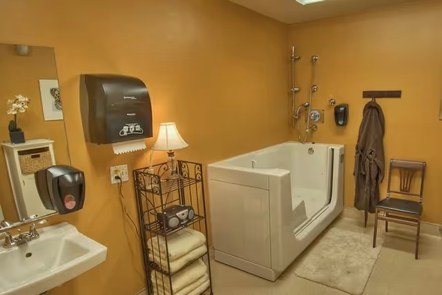 A bathroom with tan walls featuring a walk-in bathtub with a showerhead, a small white sink with a mirror above it, a paper towel dispenser, a soap dispenser, a metal rack holding folded towels and a radio, a lamp on the rack, a brown robe hanging on a wall hook, and a wooden chair.