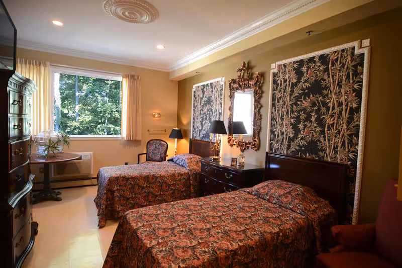 A cozy bedroom with two single beds covered in matching floral bedspreads. The room features a large window with cream curtains letting in natural light, a wooden dresser with a decorative mirror above it, two black lamps, and a patterned armchair. The walls are adorned with large framed fabric art pieces with floral and bamboo designs.