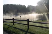 Morning mist rising over a pond with a wooden split-rail fence and trees in the background.