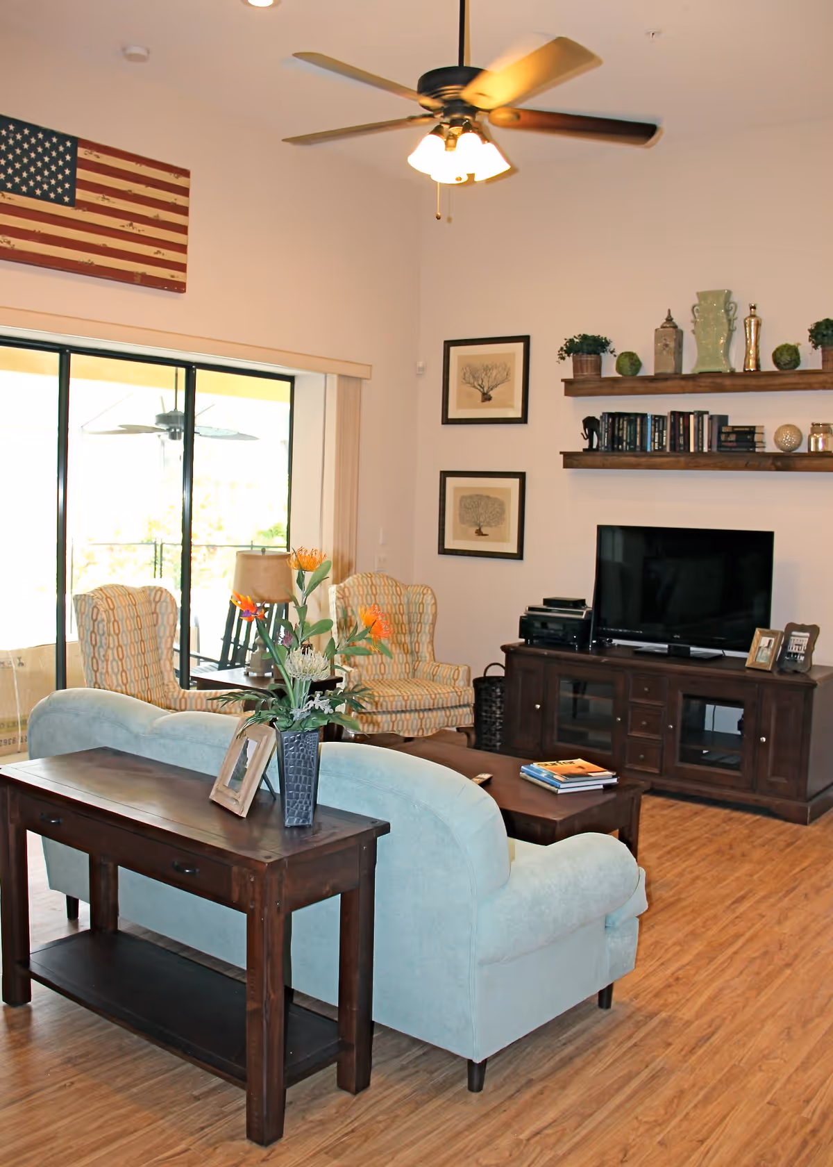 Comfortable living room with sofas, patterned armchairs, a TV console, wall shelves, and a ceiling fan.