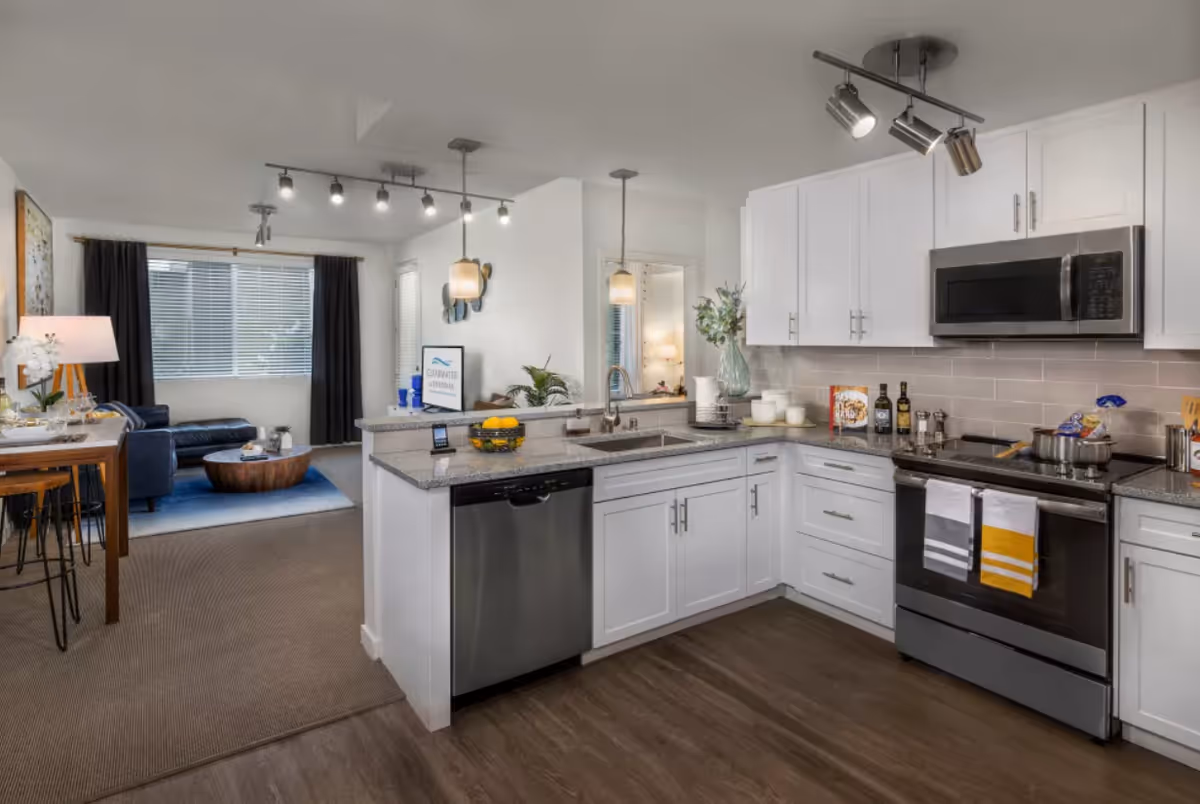 Modern kitchen with white cabinets, stainless steel appliances including a dishwasher, oven, and microwave. The kitchen has a granite countertop with a sink and decorative items like a bowl of lemons and a vase with flowers. In the background, there is a living area with a black leather sofa, a wooden coffee table on a blue rug, a window with blinds and dark curtains, and a small dining table with chairs and a lamp.