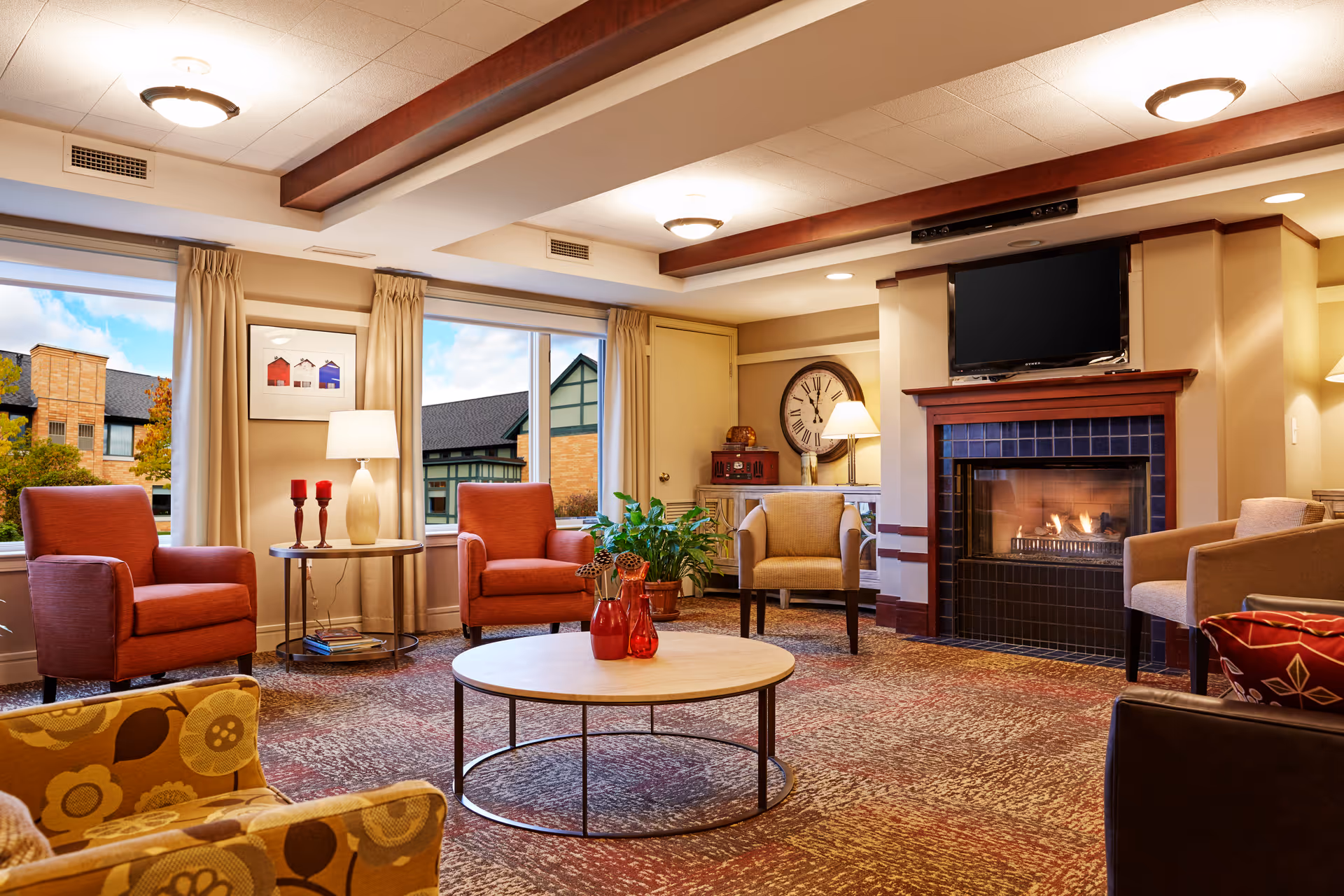 A cozy living room in Wolk Manor Assisted Living featuring a lit fireplace with a TV mounted above it, several armchairs in warm tones arranged around a round coffee table with decorative vases, large windows with beige curtains showing an outside view of buildings, a floor lamp, a large wall clock, and a patterned carpet.