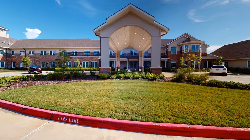 Front exterior view of Barclay House of Baton Rouge, a two-story brick and beige building with a covered entrance, surrounded by a well-maintained lawn and landscaping under a clear blue sky.