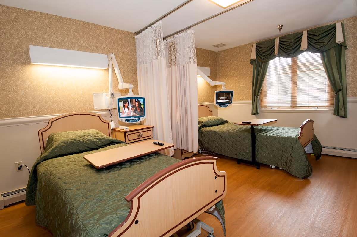 A room with two hospital-style beds separated by a privacy curtain. Each bed has a green quilted blanket, an overbed table, and a wall-mounted television. The room has wood flooring, wallpapered walls, and a window with green curtains and blinds.