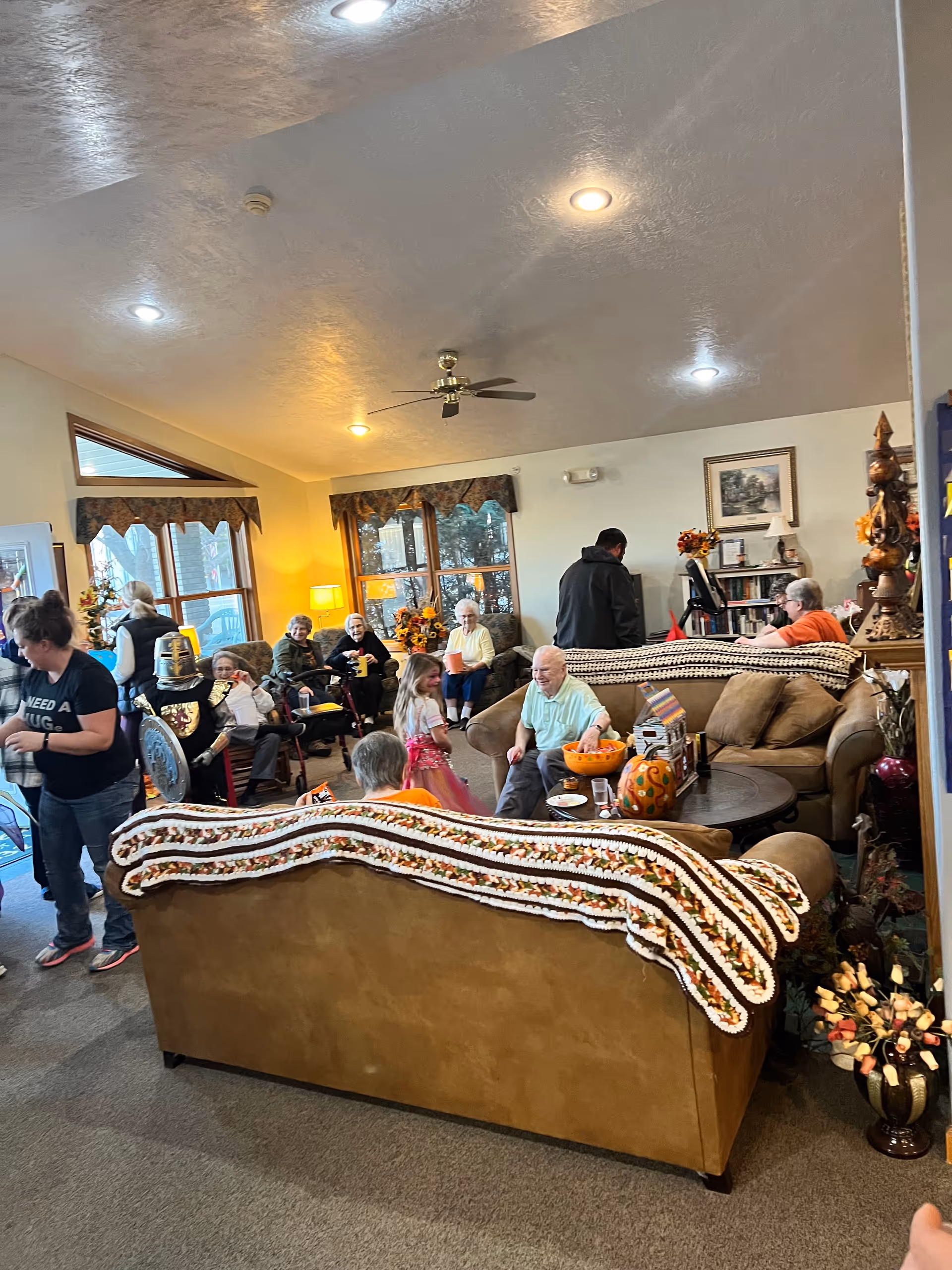 A cozy living room in a senior living facility with several elderly people and a child interacting. The room has large windows with valances, two brown sofas with crocheted blankets draped over the backs, a round coffee table with a pumpkin decoration, and various autumn-themed decorations. There is a ceiling fan and several ceiling lights illuminating the space.