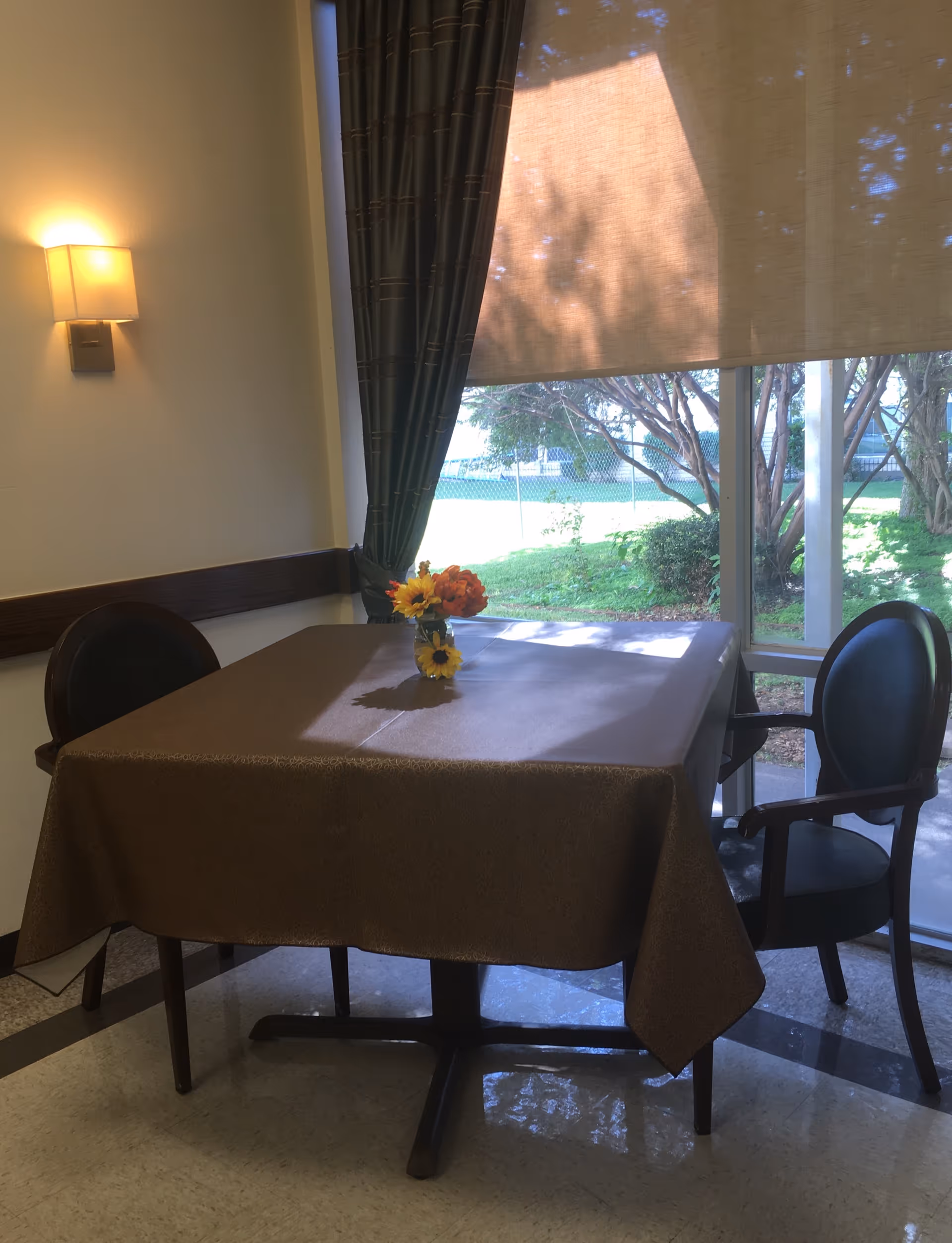 A small dining area with a square table covered by a brown tablecloth, two black chairs on opposite sides, a small vase with colorful flowers on the table, a wall-mounted light fixture, and a window with a partially drawn beige roller shade and dark curtains showing a green outdoor area.