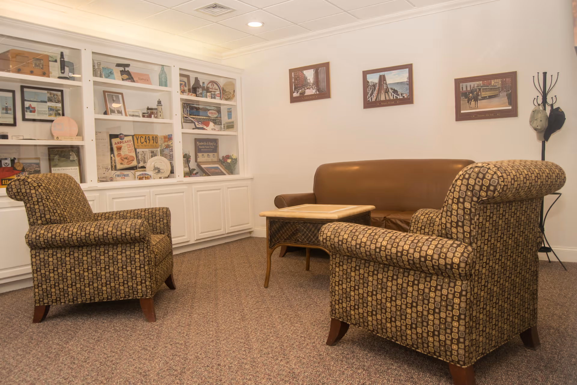 A cozy sitting area with two patterned armchairs and a brown leather sofa arranged around a wooden coffee table. Behind the seating is a white built-in cabinet with glass doors displaying various decorative items and memorabilia. Three framed pictures hang on the wall above the sofa, and a coat rack with hats is in the corner.