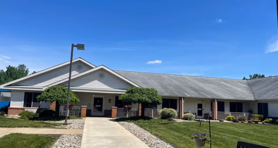 Exterior view of a single-story assisted living facility building with a gray roof, white siding, and brick accents. There is a concrete walkway leading to the main entrance, flanked by small trees and landscaped bushes. The sky is clear and blue.