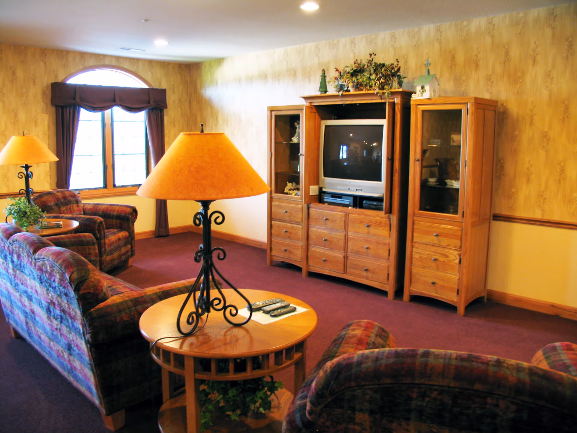 A cozy living room with patterned upholstered sofas and armchairs, a wooden entertainment center with a TV, and two round wooden side tables each holding a lamp with an orange lampshade. The room has a window with dark curtains and yellow patterned wallpaper.