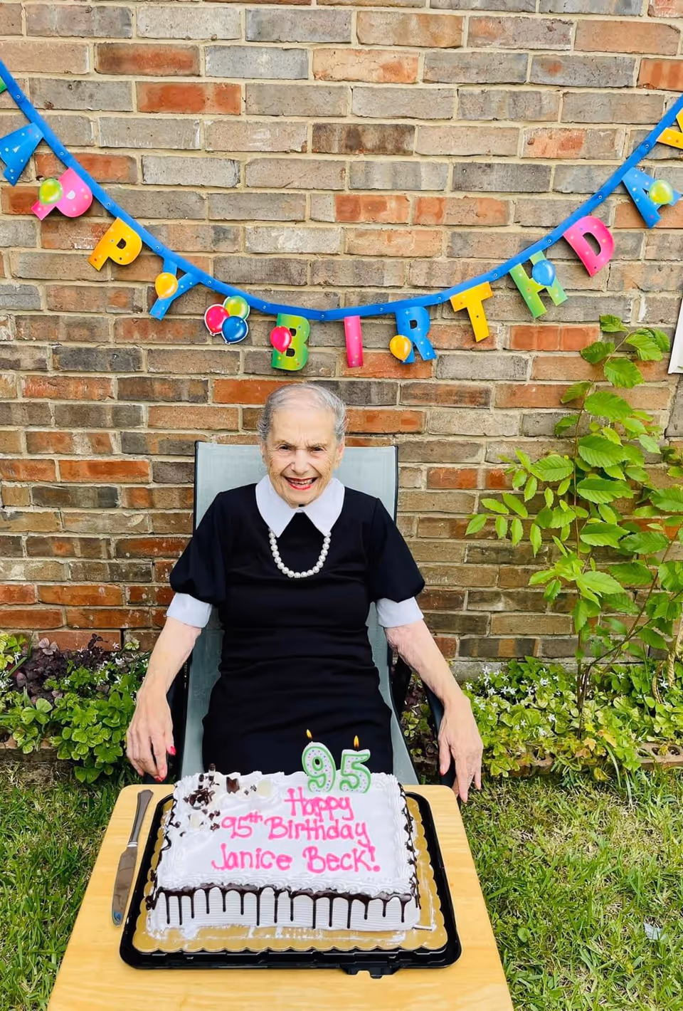 An elderly woman sitting outdoors in front of a brick wall with a colorful 'Happy Birthday' banner hanging above her. She is smiling and wearing a black dress with a white collar and a pearl necklace. In front of her on a table is a birthday cake with lit candles shaped as the number 95 and the message 'Happy 95th Birthday Janice Beck!' written in pink icing.