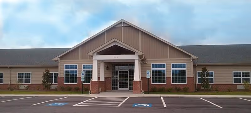 Front exterior view of Tyrrell House, a single-story building with a peaked roof, large windows, and a covered entrance. The parking lot in front includes marked handicap parking spaces.