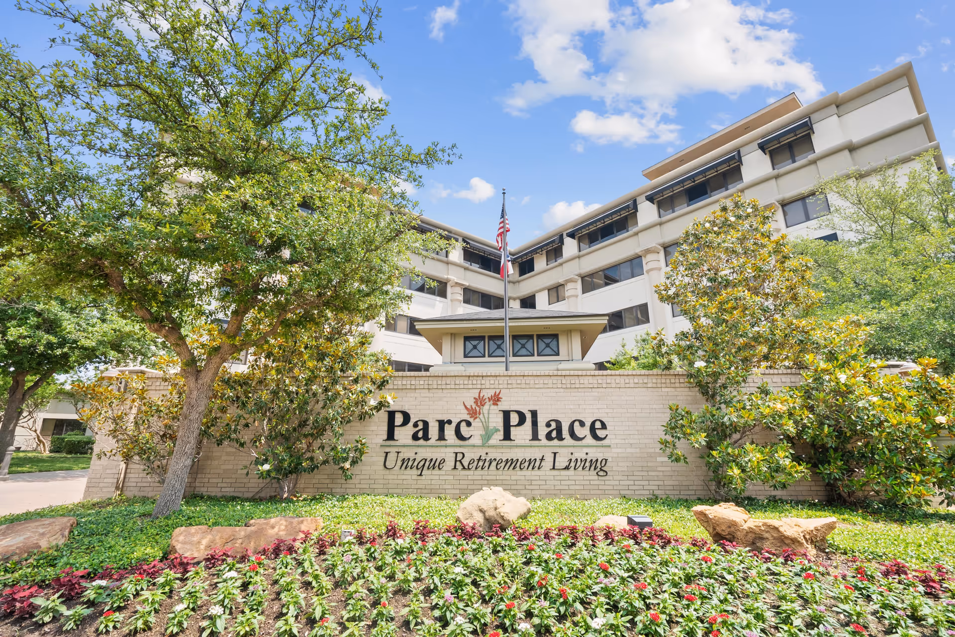 Exterior view of Parc Place retirement living facility with a large sign displaying the name and slogan, surrounded by trees, bushes, and a flower bed under a partly cloudy sky.