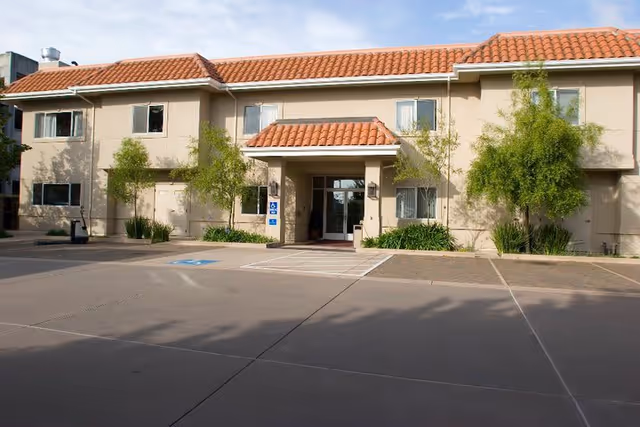 Exterior view of a two-story building with a tiled roof and beige walls, featuring a covered entrance with glass doors. There are small trees and shrubs planted along the front, and a parking area with marked spaces, including a handicap parking spot, in the foreground.
