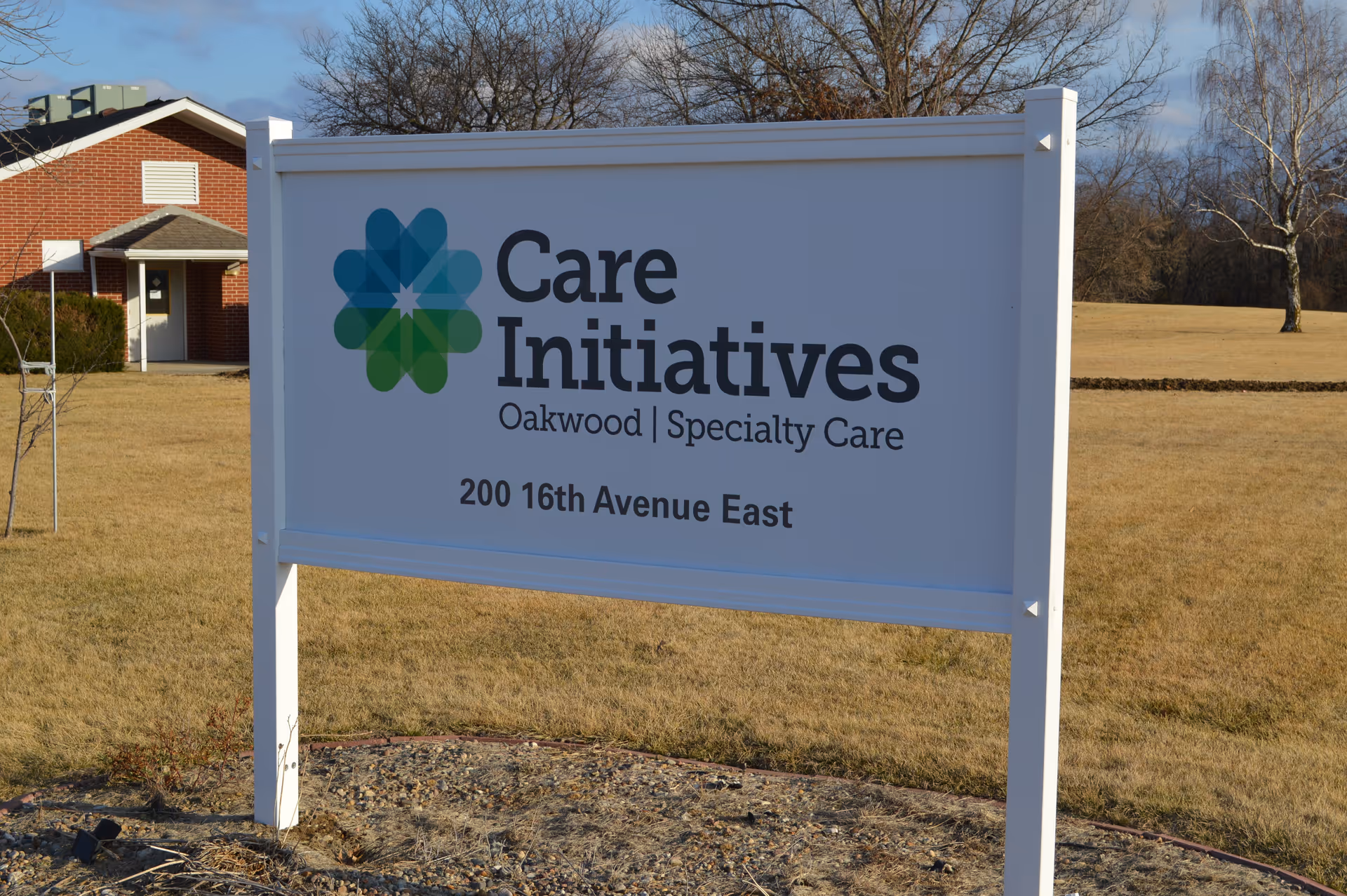 A white freestanding sign reading 'Care Initiatives Oakwood | Specialty Care 200 16th Avenue East' stands on a grassy lawn with a brick building and trees in the background.