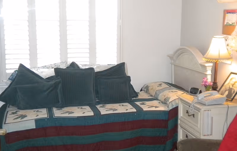 A cozy bedroom with a bed covered in a quilted blanket featuring a nature-themed pattern and several dark green pillows. Next to the bed is a white nightstand with a telephone, a small lamp with a beige shade, a framed photograph, and a small vase with pink flowers. White plantation shutters cover the window behind the bed.