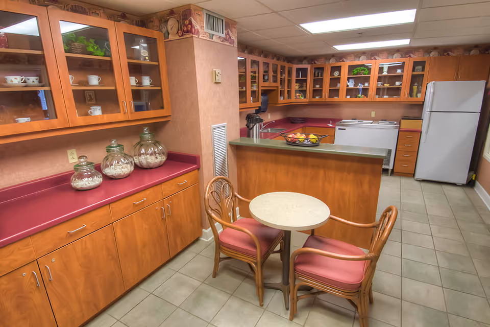 Communal kitchen area with wood cabinets and glass-front cupboards, a countertop island, refrigerator, and a small round table with two chairs.