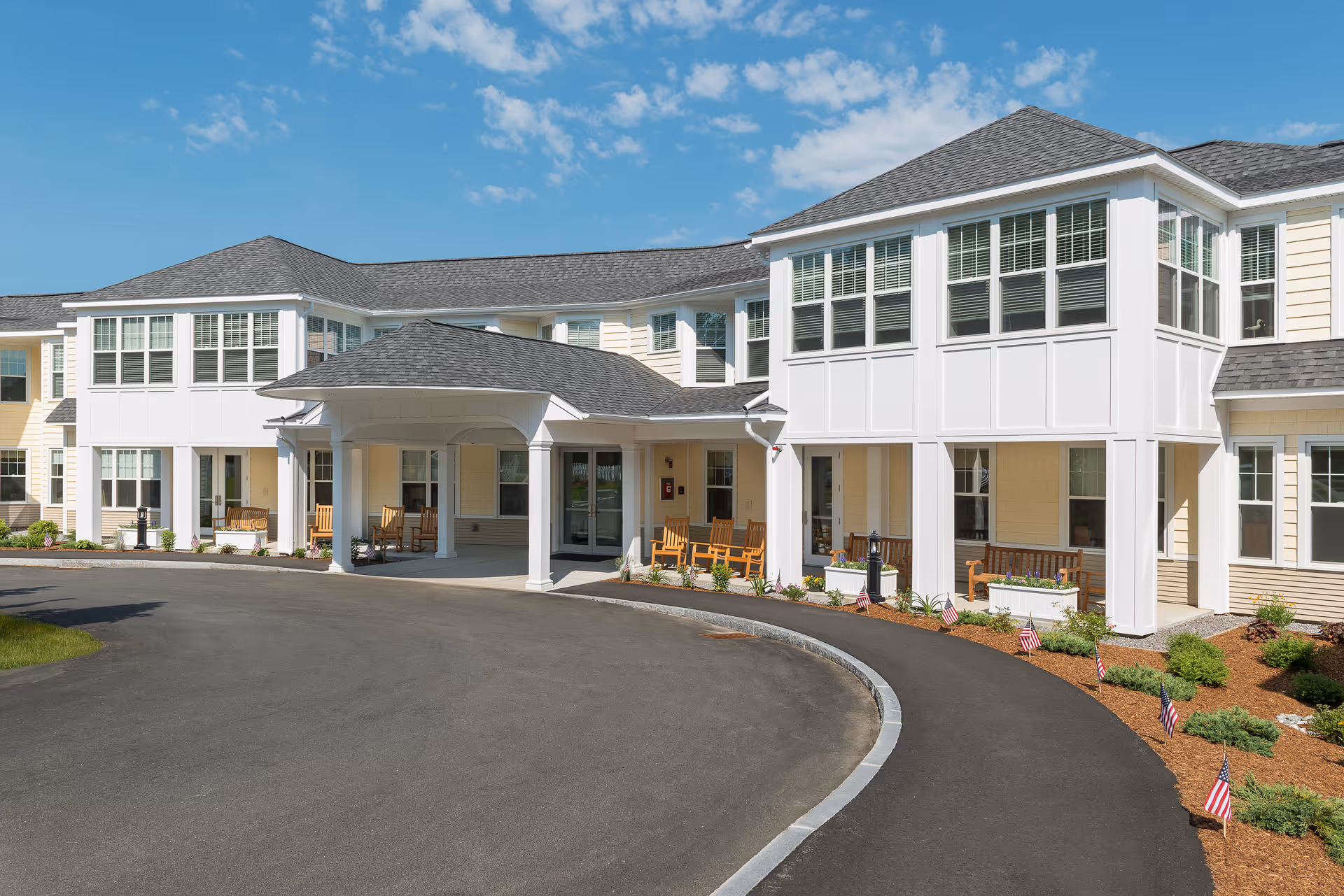 Front exterior of a two-story senior living facility with a covered entrance, circular driveway, benches, and small American flags lining the landscaping.