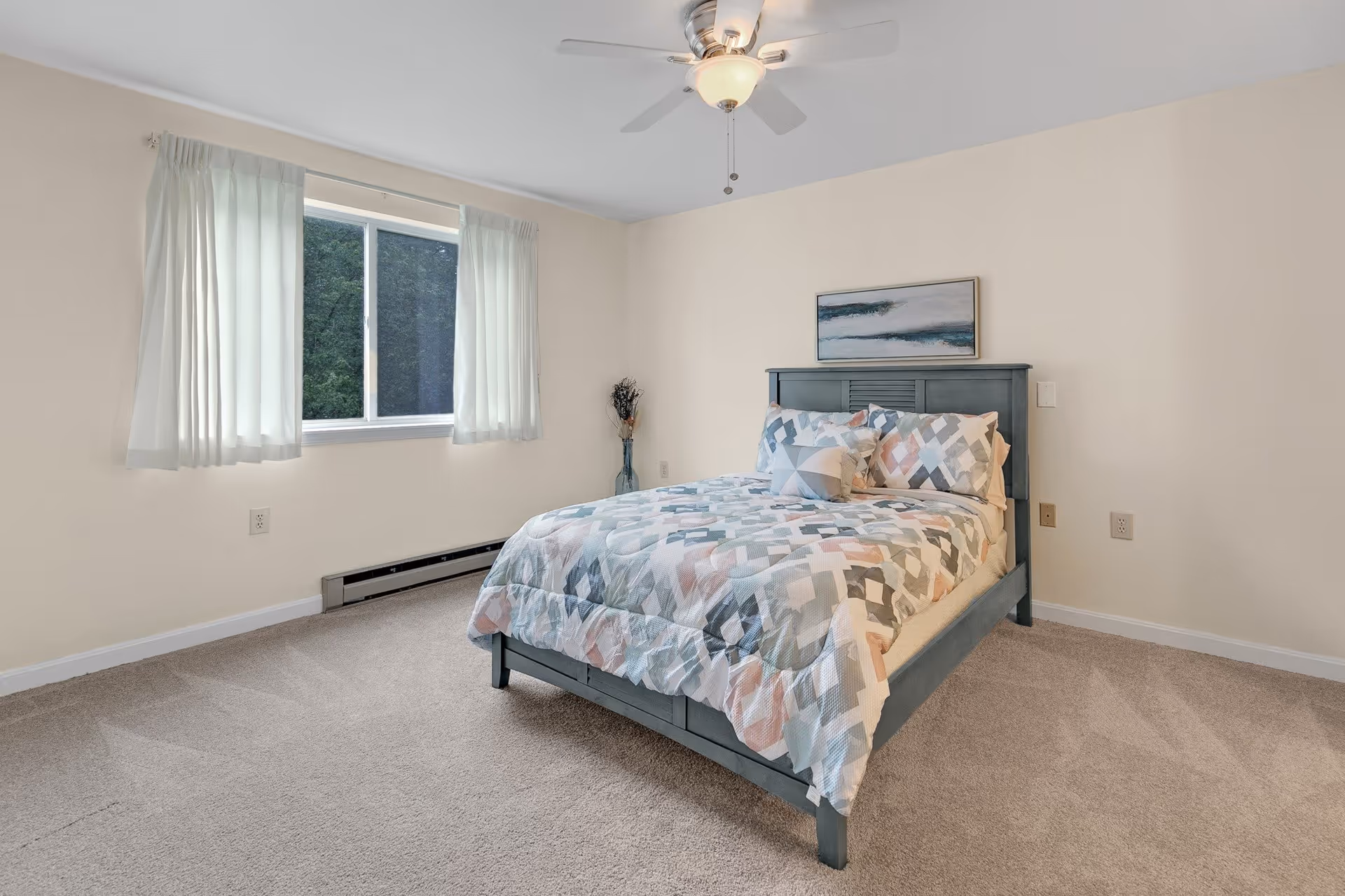 A simple bedroom with a double bed featuring a patterned comforter and pillows. The room has beige walls, a window with sheer white curtains, a ceiling fan with a light, and a framed abstract painting above the bed. The floor is carpeted in a neutral tone.