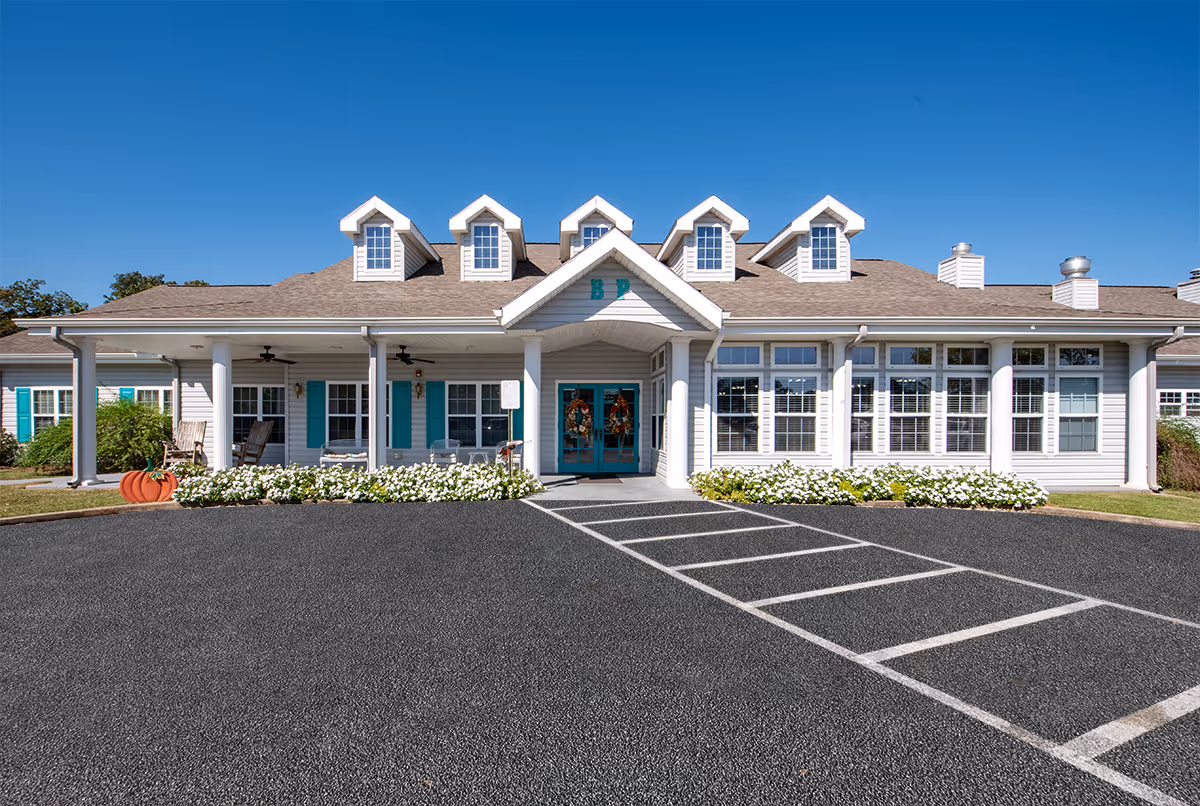 White senior living facility with teal doors and flower beds in front of a parking lot.