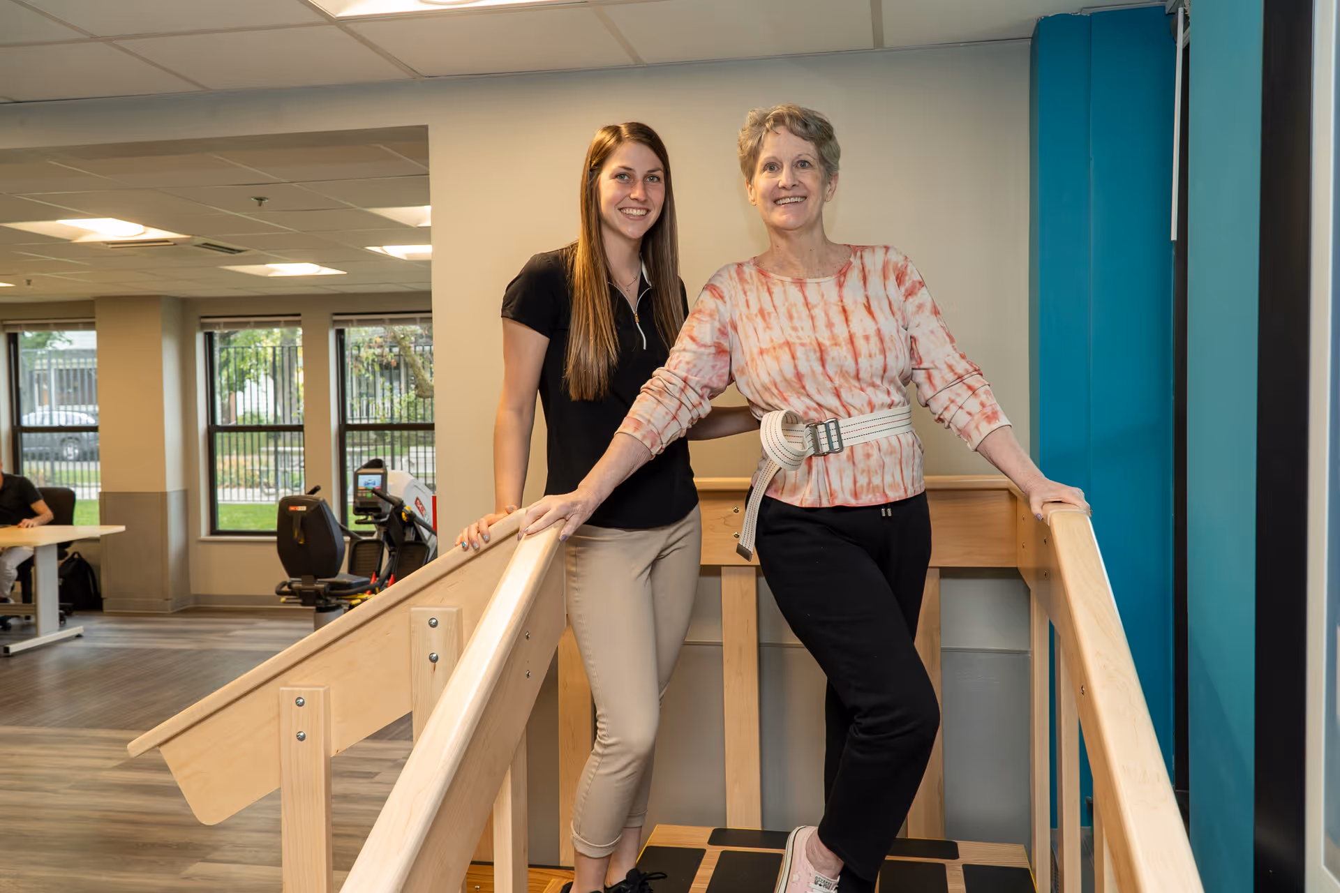 A young female therapist assists an older woman who is using parallel bars for walking exercises in a rehabilitation center. The older woman is wearing a gait belt and smiling, while the therapist stands beside her, also smiling. The background shows exercise equipment and large windows with a view outside.