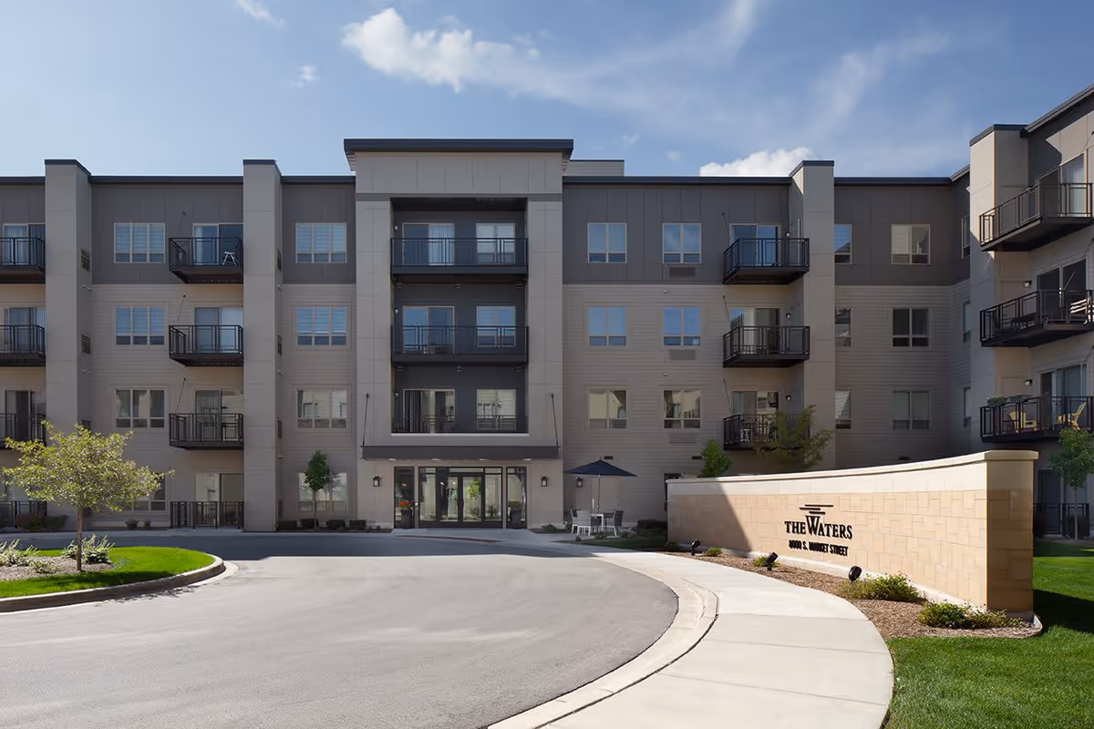Front exterior view of The Waters of Oak Creek senior living facility, showing a modern multi-story building with balconies, a curved driveway, landscaped greenery, and a sign with the facility's name and address.