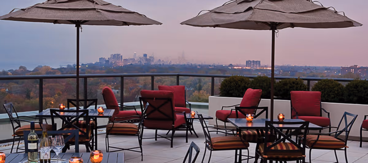 Outdoor patio area with tables, chairs, and umbrellas overlooking a distant city skyline at dusk. The seating includes cushioned chairs with red upholstery and small candle lights on the tables.