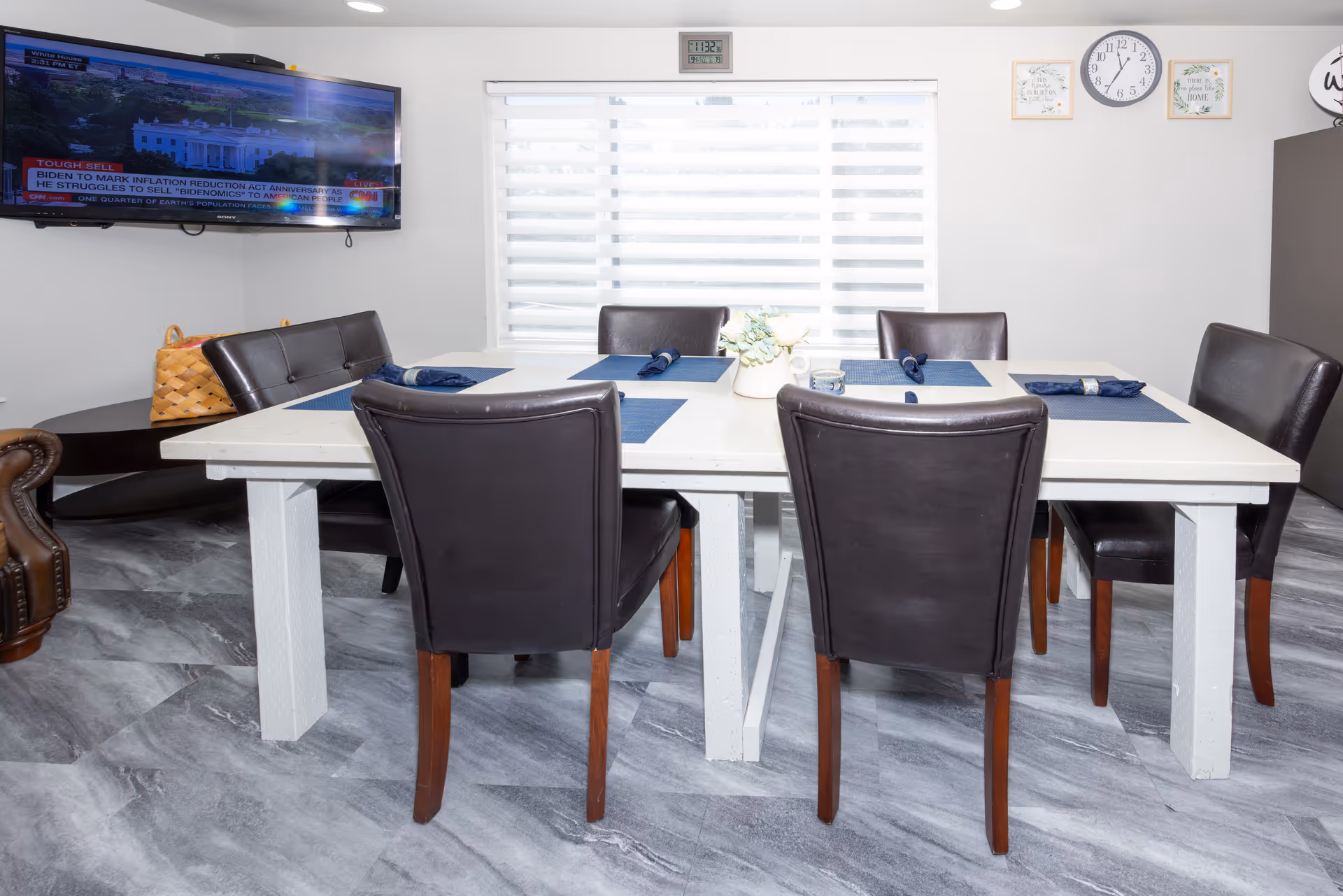 A dining room with a white rectangular table set with six dark brown leather chairs. The table has blue placemats and rolled blue napkins. A small plant centerpiece is on the table. A large window with blinds is behind the table, and a wall clock and framed decor hang above the window. A flat-screen TV is mounted on the left wall, showing a news channel. The floor has gray marble-like tiles.