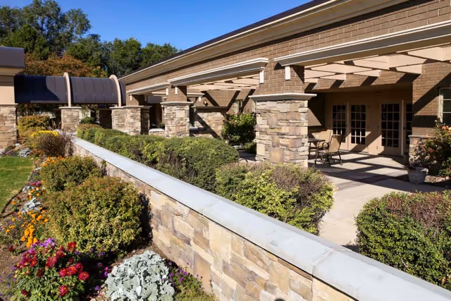 Outdoor patio area of a building with stone pillars and a pergola, surrounded by well-maintained bushes and colorful flowers along a stone wall under a clear blue sky.