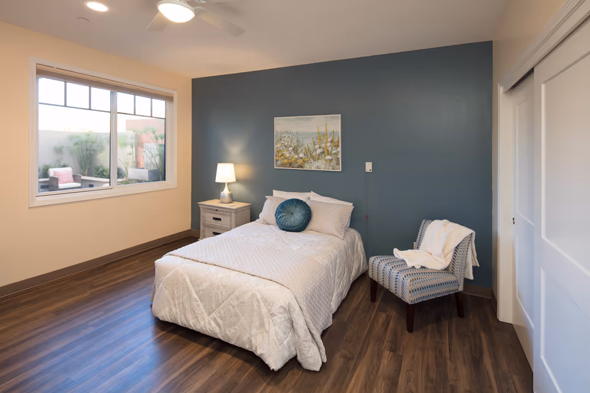 A cozy bedroom with a bed covered in a white quilt and pillows, including a round blue decorative pillow. There is a nightstand with a lamp on the left side of the bed, a blue accent wall behind the bed with a framed floral painting, and a patterned chair with a white blanket draped over it on the right. The room has wooden flooring, a large window showing an outdoor seating area with plants, and white sliding closet doors.