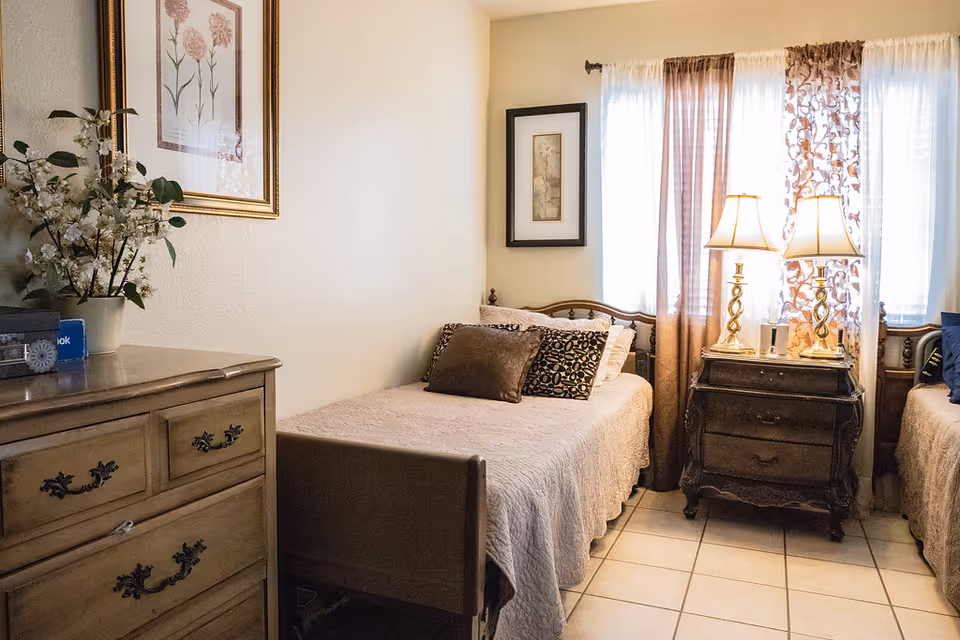A cozy bedroom with two single beds separated by a dark wooden nightstand with two lamps. The beds have beige bedspreads and decorative pillows. There is a wooden dresser with a potted plant and a book on top. The room has tiled flooring, light-colored walls, and windows with sheer and patterned curtains allowing natural light to enter.