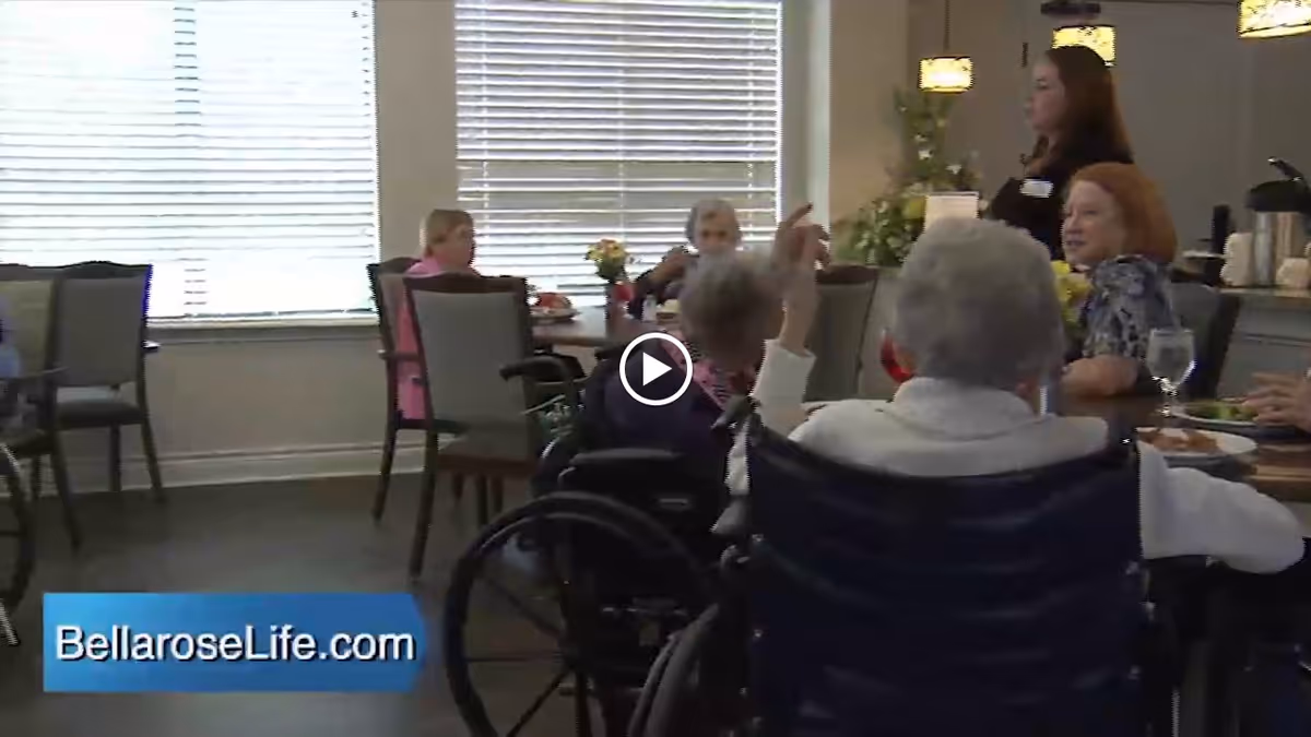 Several elderly individuals seated around tables in a dining area, with a caregiver standing nearby. The room has large windows with blinds and is well-lit.