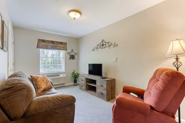 A cozy living room with a brown sofa on the left and a red recliner chair on the right. A wooden TV stand with a flat-screen television is against the far wall, which is decorated with a metal wall art piece. A window with a patterned valance lets in natural light, and a floor lamp stands next to the recliner.
