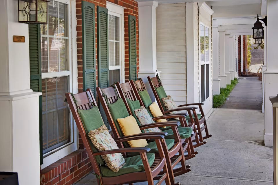 A row of wooden rocking chairs with green cushions and decorative pillows lined up on a covered porch outside a building with brick and white siding walls, green shutters on the windows, and a hanging lantern light fixture.