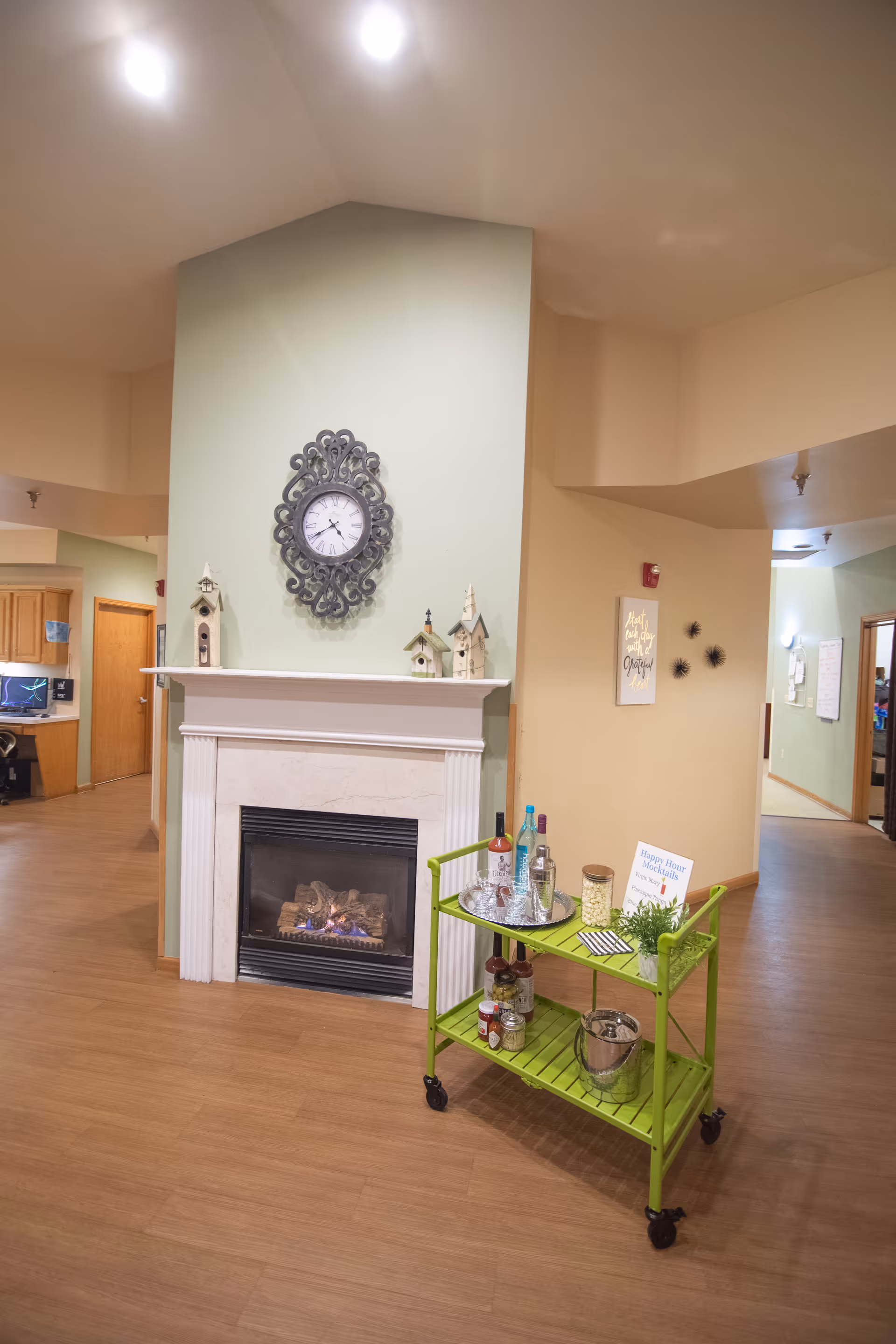 Interior view of a common area in an assisted living facility featuring a lit fireplace with a decorative clock and birdhouse figurines on the mantel. In front of the fireplace is a green rolling cart with bottles, glasses, and snacks. The room has wood flooring and light-colored walls with some wall decorations.