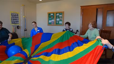 A group of elderly individuals and a caregiver sitting in a circle indoors, holding the edges of a colorful parachute with blue balls on top of it, engaging in a group activity.