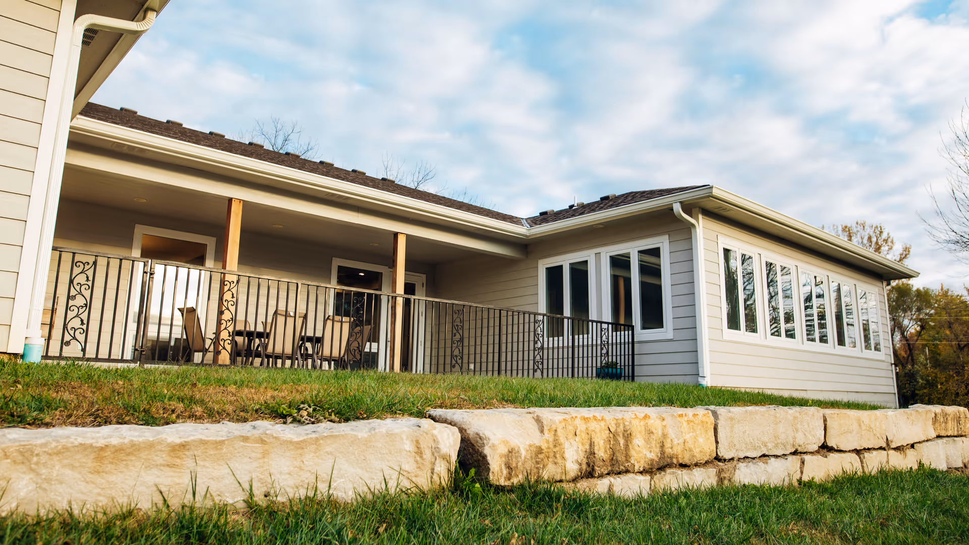 Exterior view of a single-story building with a covered porch featuring outdoor seating and a railing, surrounded by grass and stone landscaping under a partly cloudy sky.