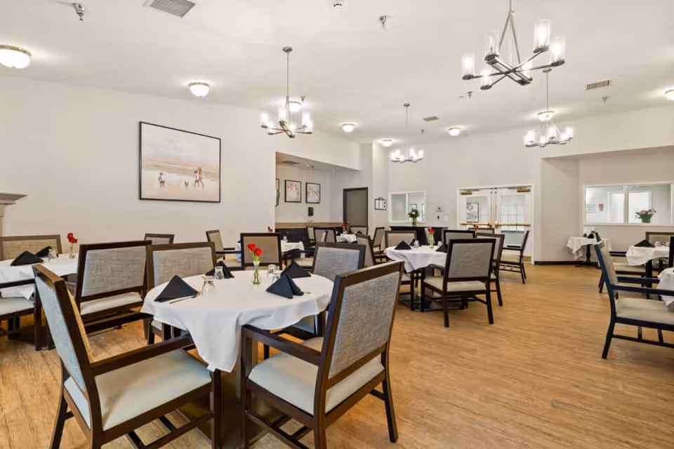 A bright and spacious dining room with multiple round tables covered with white tablecloths, each set with black napkins, glasses, and small vases with red flowers. The room has wooden flooring, modern chandeliers, and light-colored walls adorned with framed artwork. There are several chairs around each table, and the room appears clean and well-organized.