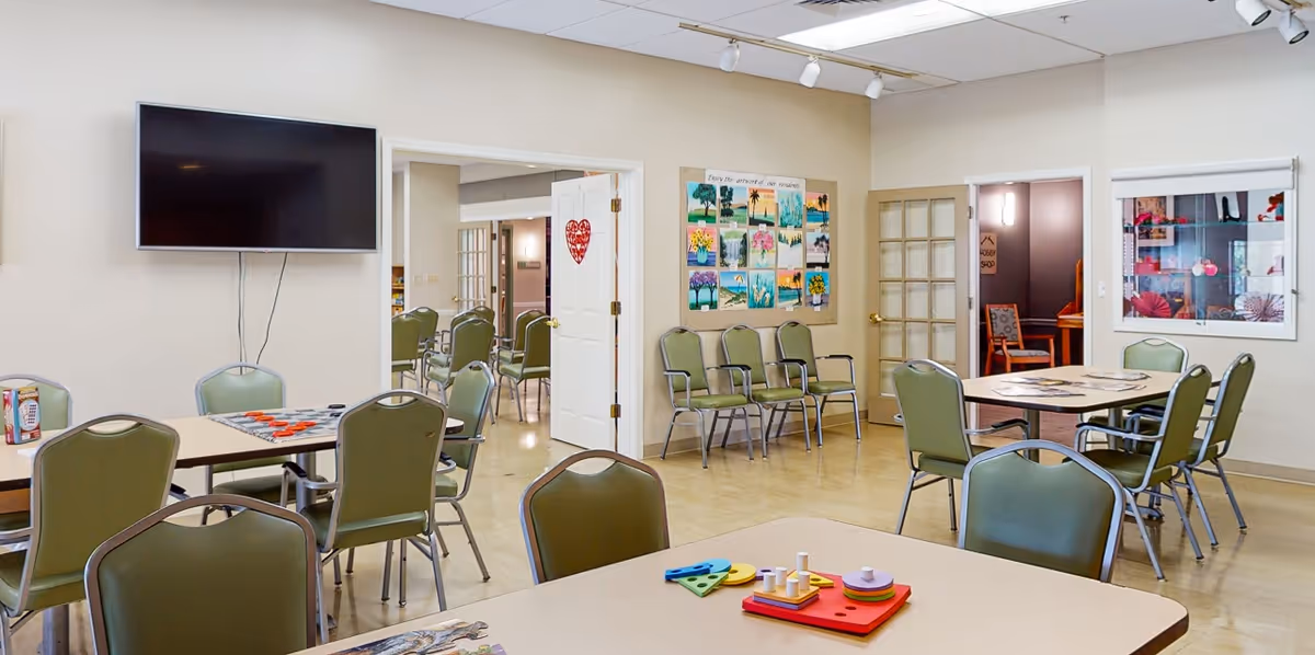 A bright activity room with several tables and green chairs arranged around them. On the tables are various colorful puzzles and games. A large flat-screen TV is mounted on the wall. The walls are decorated with a bulletin board displaying artwork and a heart decoration on a white door. There are open doorways leading to other rooms, and a display window showing crafts or decorations.
