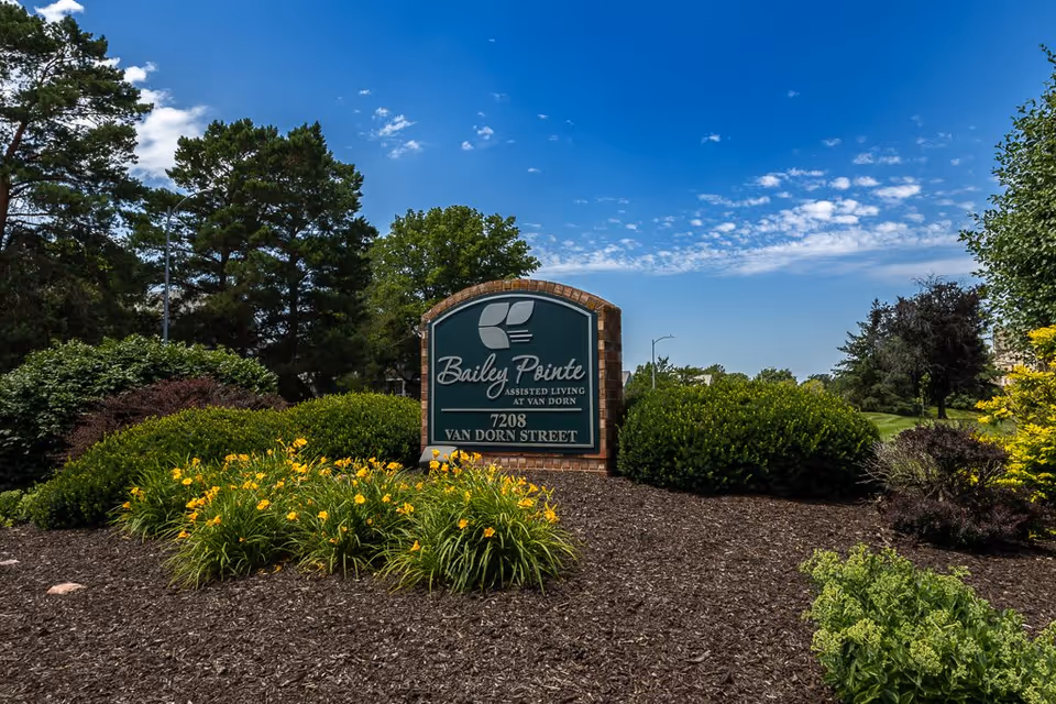 Outdoor landscaped area with bushes, yellow flowers, and trees surrounding a green and brick sign that reads 'Bailey Pointe Assisted Living at Van Dorn, 7208 Van Dorn Street' under a partly cloudy blue sky.