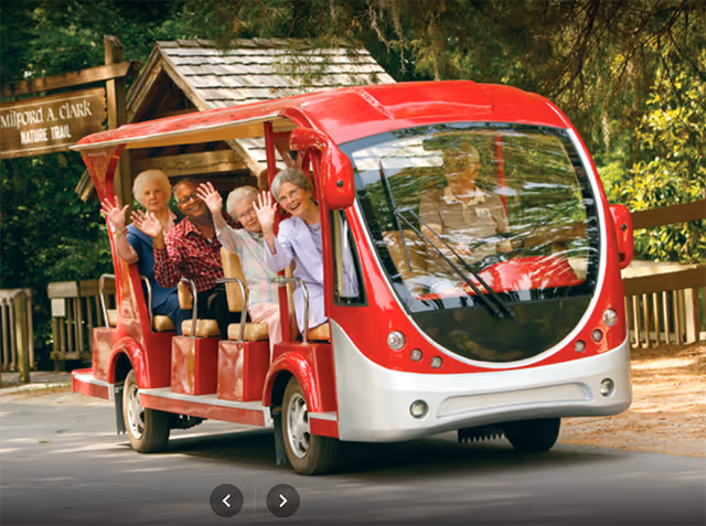 A group of elderly people happily waving while riding in a red and white open-sided electric shuttle vehicle on a paved path surrounded by trees and greenery near a wooden sign that reads 'Milford A. Clark Nature Trail.'