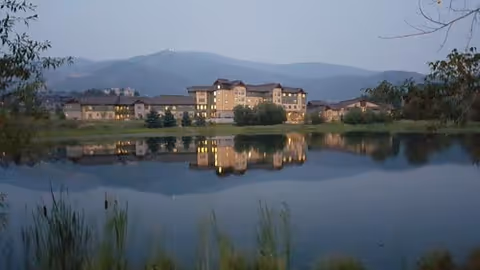 A large building complex reflected in a calm body of water with mountains in the background during dusk or early evening.